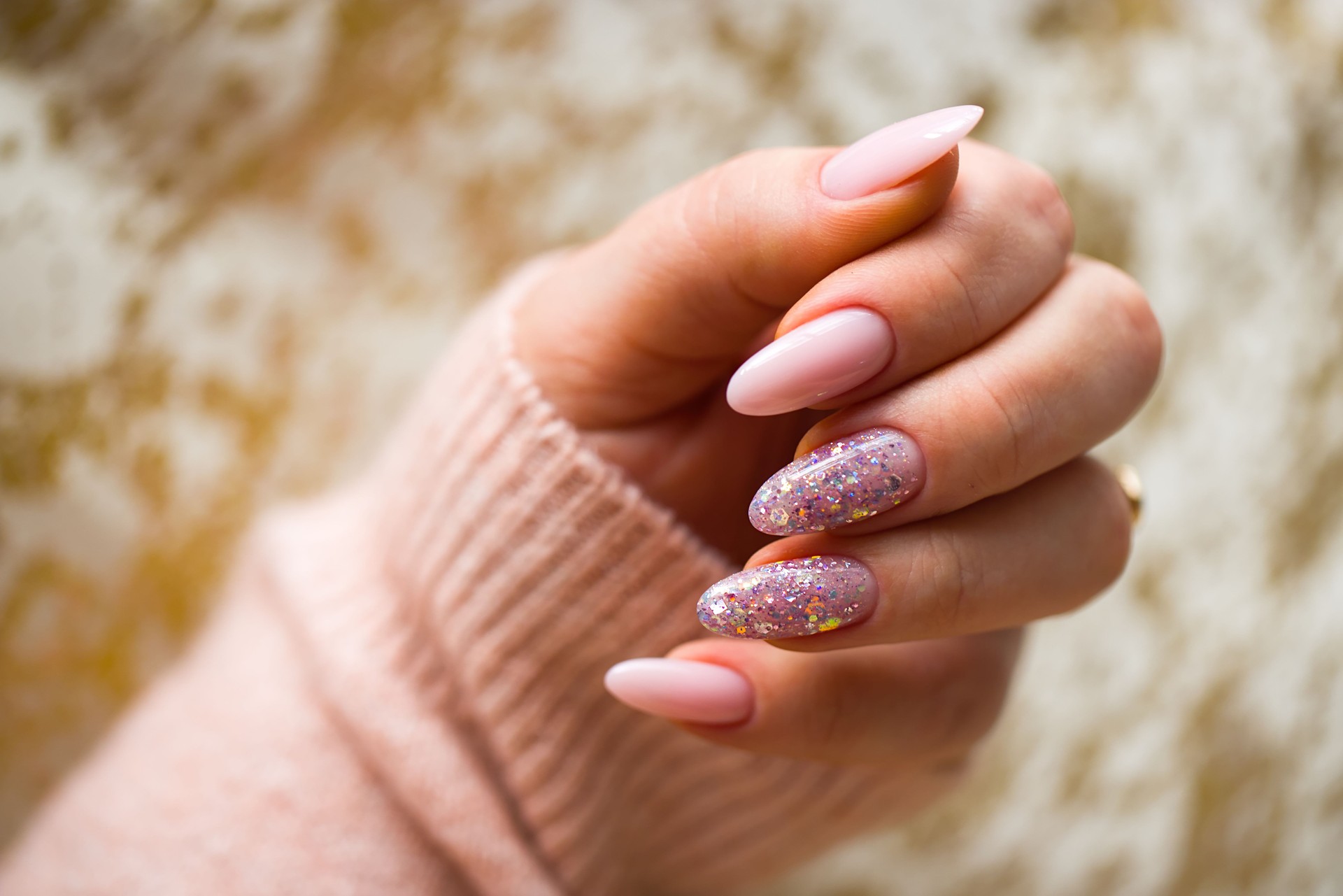 A photo of a woman's hand with beautiful pink gel nails. The hand is well-groomed, and the nails are perfectly painted, with two adorned with stylish, multicolored glitter.